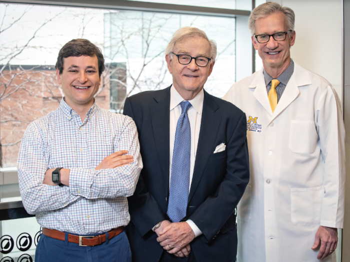 Three white men are standing in front of a window. They are facing forward and smiling.