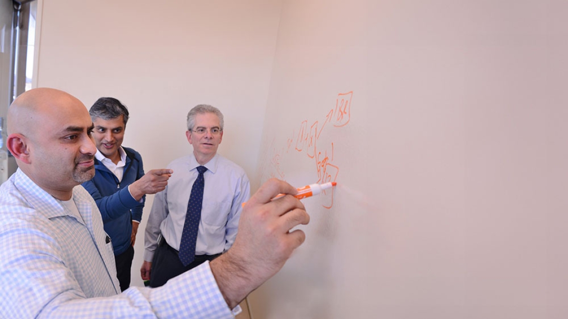 Three faculty working together at a whiteboard. One is writing with an orange marker as the other two are watching.